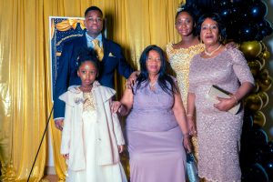 A family poses together in formal attire against a backdrop of gold and black decorations, celebrating a special occasion. The group includes three women and two children, all smiling.