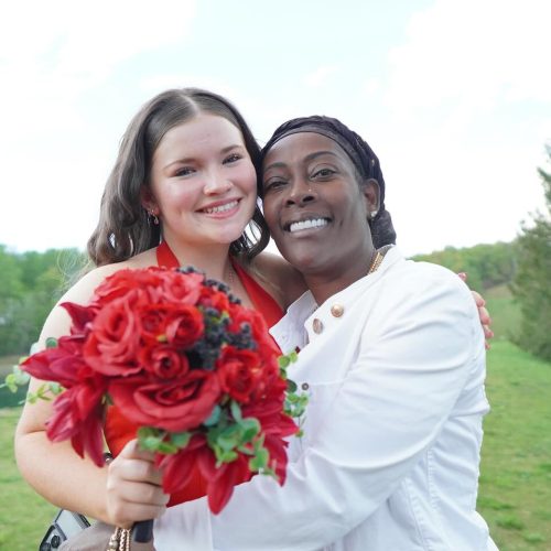 Two women embrace joyfully outdoors, with one holding a bouquet of red roses. The background features greenery and a body of water, suggesting a celebratory occasion.