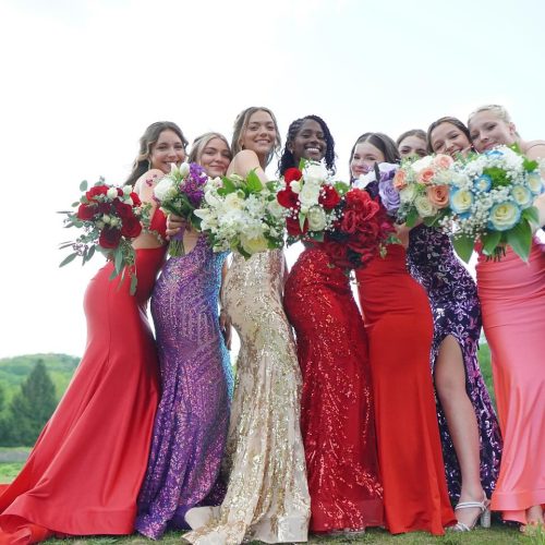 A group of nine women in colorful evening gowns stands together, each holding a bouquet of flowers, smiling and posing outdoors. The scene captures a festive and joyful atmosphere.