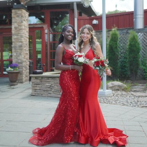 Two women in elegant red dresses pose together outdoors, smiling and holding a bouquet. The background features a rustic building and greenery.