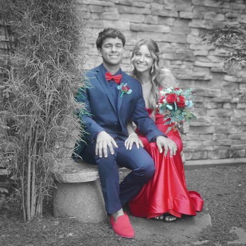 A young couple in red and blue formal attire sits joyfully on a stone bench against a brick wall, surrounded by lush greenery. The woman holds a vibrant bouquet.
