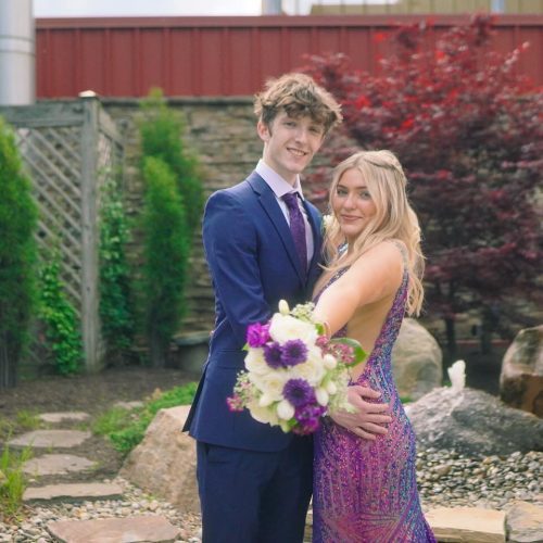 A young couple poses together in a garden setting, dressed elegantly for a special occasion. The woman holds a bouquet of flowers, and they both smile happily.