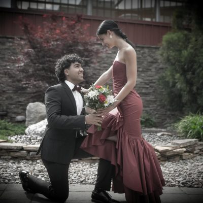 A man kneels with a bouquet, presenting it to a woman in an elegant red dress, surrounded by a garden setting. The scene conveys a romantic moment.