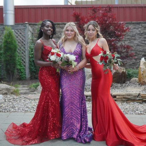 Three young women pose together outdoors, each wearing elegant evening gowns in vibrant colors, holding bouquets of flowers. The setting features greenery and decorative stones in the background.