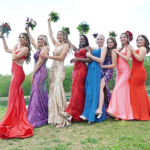 A group of eight women in colorful evening gowns poses together outdoors, each holding a bouquet. They display joyful expressions, celebrating a special occasion.