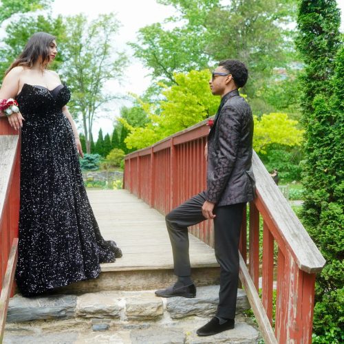 A couple dressed elegantly stands on a wooden bridge surrounded by greenery, with the woman in a black gown and the man in a stylish suit. The scene conveys a romantic and formal atmosphere.