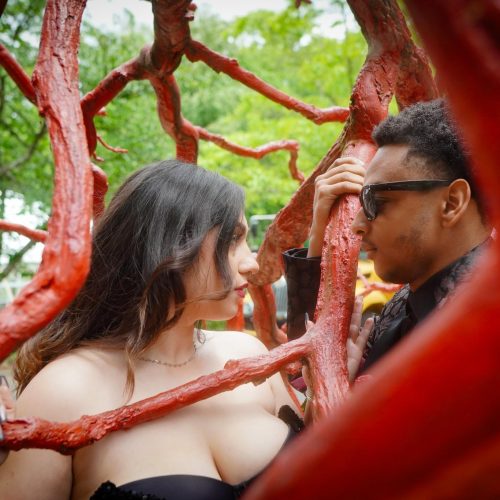 A couple interacts playfully within a vibrant, red tree sculpture in a lush outdoor setting. The scene captures a moment of connection amidst the unique artistic structure.