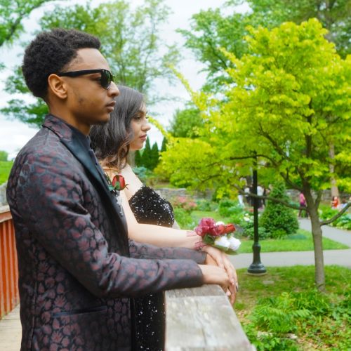 A young man in a stylish black suit and sunglasses stands beside a woman in a formal dress, both gazing thoughtfully into the distance. They are surrounded by lush greenery in a park setting.