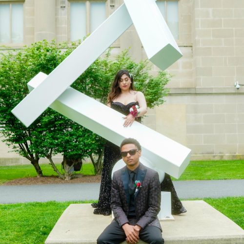 A young man in a suit sits in front of a modern geometric sculpture, while a woman in a black dress stands behind it. They pose confidently in front of a stately building.