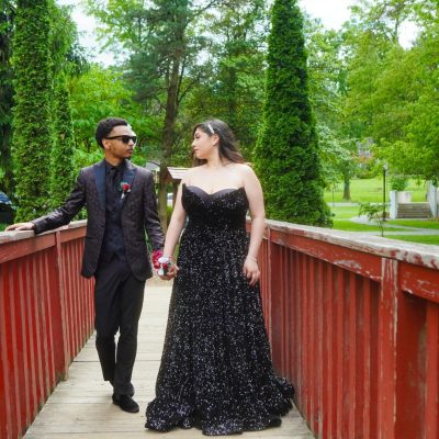 A couple walks hand in hand across a wooden bridge surrounded by lush greenery, with the woman wearing an elegant black gown and the man dressed in a formal suit. The scene captures a moment of connection and celebration.