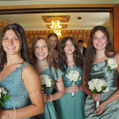 Four young women in teal dresses stand together, each holding a bouquet of white roses, smiling as they pose in a warmly lit room. The atmosphere suggests a celebratory occasion, likely a wedding or formal event.