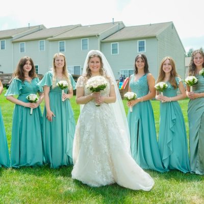 A bride stands in the center, surrounded by her bridesmaids, all wearing teal dresses and holding bouquets. The setting is a grassy area with houses in the background.