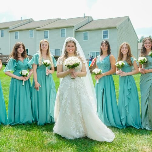 A bride stands in the center, surrounded by her bridesmaids, all wearing teal dresses and holding bouquets. The setting is a grassy area with houses in the background.
