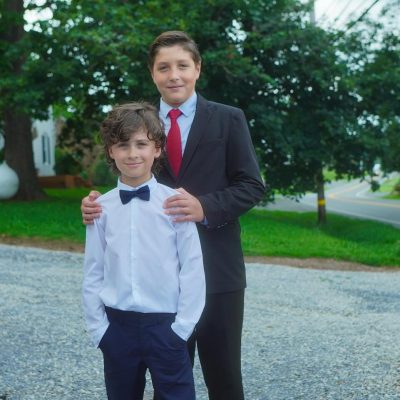 Two boys dressed in formal attire stand together outdoors, with one boy in a white shirt and bow tie and the other in a suit and tie. The background features greenery and a gravel path.