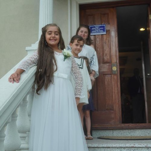 A young girl in a white dress smiles on a staircase outdoors. Two other children stand behind her at a wooden door. The scene is joyful and bright.