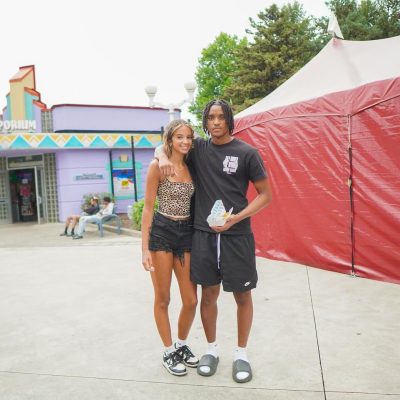 A young couple poses together in a colorful amusement park setting, with a vibrant building and a large tent in the background. They both appear cheerful and relaxed.