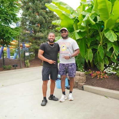Two men stand together in a lush outdoor setting, surrounded by green plants and colorful flowers. They are smiling and dressed casually, enjoying a sunny day.