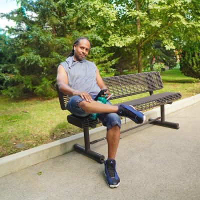A man is sitting on a park bench, casually checking his phone while surrounded by greenery. The scene conveys a relaxed atmosphere.