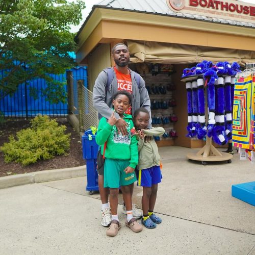 A father stands with his two children in front of a boathouse, surrounded by colorful merchandise displays. The family appears cheerful and engaged in their outing.