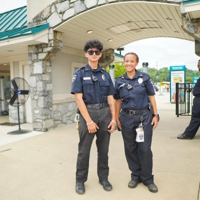 Two police officers stand together at the entrance of a park, smiling and dressed in their uniforms. The background features an archway and a sign for the park.