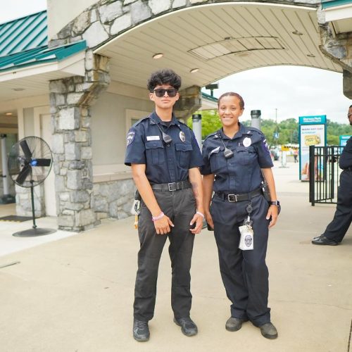 Two police officers stand together at the entrance of a park, smiling and dressed in their uniforms. The background features an archway and a sign for the park.