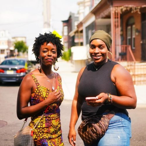 Two women are walking together on a sunny street, smiling and engaging with each other. They are dressed in colorful outfits, reflecting a vibrant and lively atmosphere.