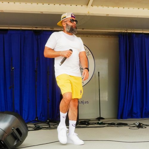 A man in a white t-shirt and yellow shorts performs on stage, holding a microphone, with a blue curtain in the background. The scene captures the energy of a live performance.