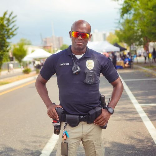 A police officer stands confidently on a street, wearing sunglasses and a uniform, with a community event taking place in the background. The scene conveys a sense of security and community engagement.