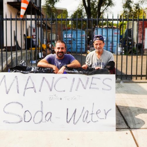 Two men sit behind a large sign that reads "MANCINES Soda Water," with a fence and some containers visible in the background. The scene appears to be a casual outdoor setup, likely for selling beverages.
