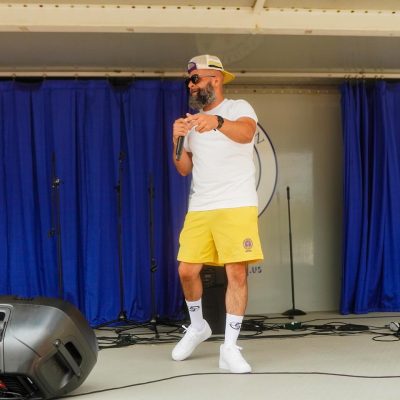 A man performs on stage, wearing a white t-shirt and yellow shorts, with a microphone in hand and a lively expression. The backdrop features blue curtains, creating a vibrant atmosphere.