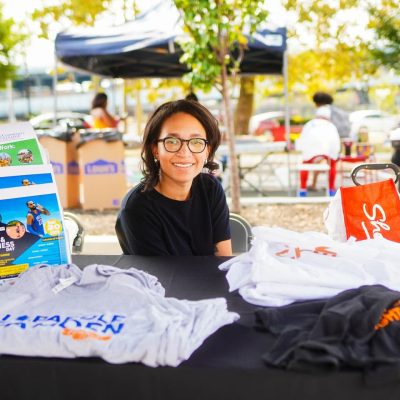 A young woman smiles while sitting behind a table displaying various T-shirts at an outdoor event. Colorful tents and people can be seen in the background, creating a lively atmosphere.