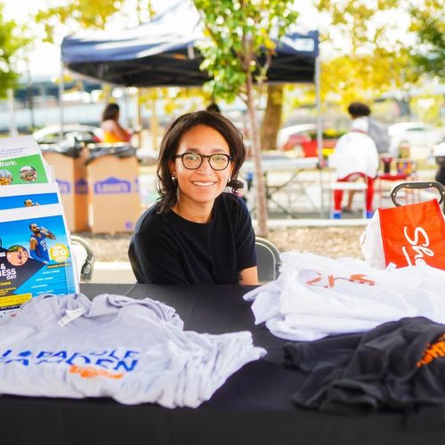 A young woman smiles while sitting behind a table displaying various T-shirts at an outdoor event. Colorful tents and people can be seen in the background, creating a lively atmosphere.