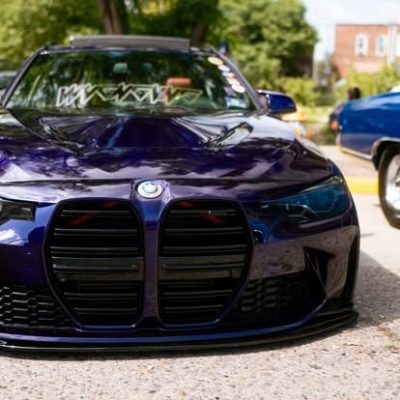 A sleek purple sports car with a distinctive front grille is parked on a sunny street, showcasing its modern design and polished finish. The background features greenery and other vehicles, adding to the vibrant atmosphere.
