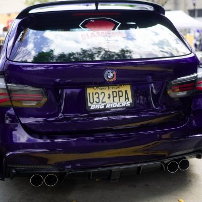 A compact car with a striking purple exterior, featuring a sporty rear design and dual exhaust pipes, is parked in a lively outdoor setting. The license plate is clearly visible.