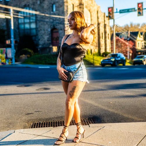 A young woman poses confidently on a street corner, wearing a stylish outfit that includes a black top and denim shorts. The background features a mix of urban elements and greenery.