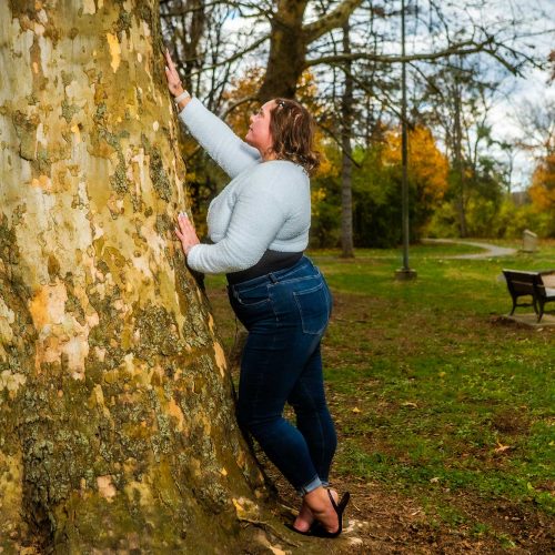 A person leans against a large tree in a park, surrounded by autumn foliage and a grassy area. A bench is visible in the background.