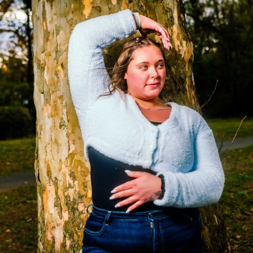 A young woman with curly hair leans against a tree, wearing a light blue cardigan and dark jeans, in a park setting. The background features blurred greenery and a pathway.