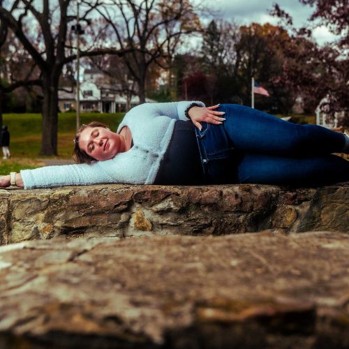 A woman lies playfully on a stone surface in a park, smiling at the camera, surrounded by trees and grassy areas. The scene conveys a relaxed and cheerful atmosphere. --- Please let me know if you need any further assistance!