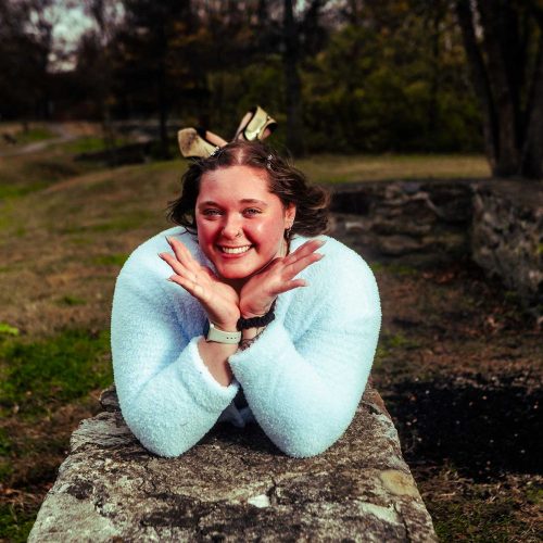 A smiling woman with a bow in her hair poses playfully while lying on a stone wall in a park setting. The background features greenery and trees, creating a cheerful atmosphere.