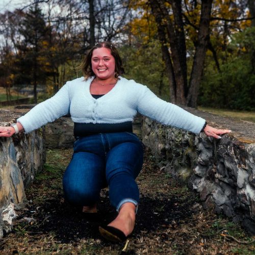 A woman is kneeling on a stone structure in a park, smiling and wearing a light blue sweater and blue jeans. The background features trees with autumn foliage.