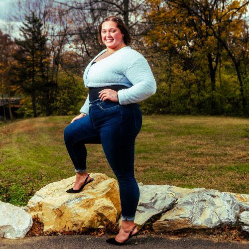 A woman stands confidently on a rock in a park, wearing a light sweater and high-waisted jeans, with trees and a cloudy sky in the background.