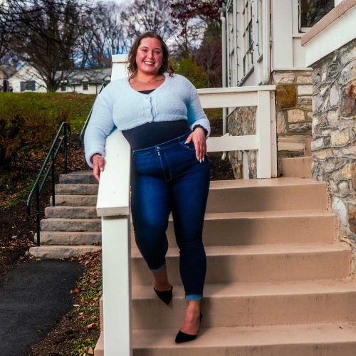 A woman walks confidently down a set of stairs, wearing a light blue top and dark jeans, with a smile on her face. The background features a grassy area and a house.