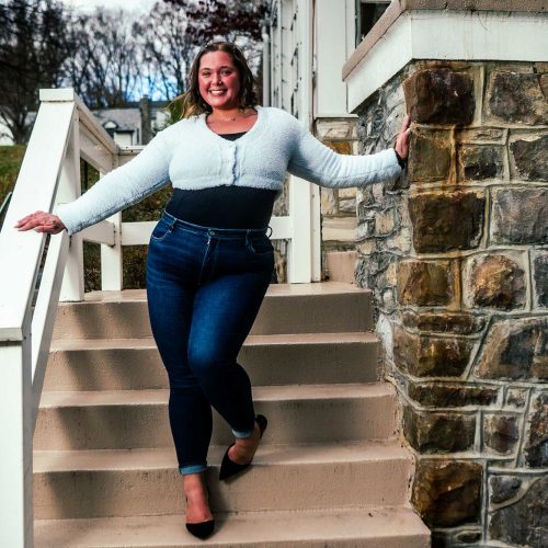 A young woman confidently walks down a set of stairs, wearing a stylish outfit that includes a light sweater and fitted jeans. The setting features a stone wall and greenery in the background.