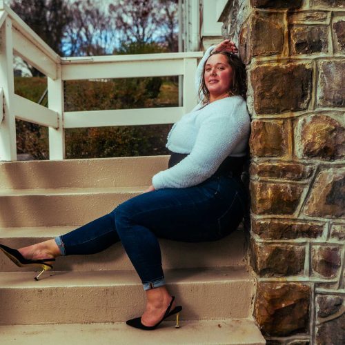 A woman in a white sweater and jeans sits confidently on outdoor steps, leaning against a stone wall. She exudes a relaxed, confident vibe.