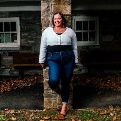 A young woman stands confidently against a stone pillar, wearing a white top and blue jeans, with autumn leaves scattered on the ground around her. The background features a rustic building with windows and benches.