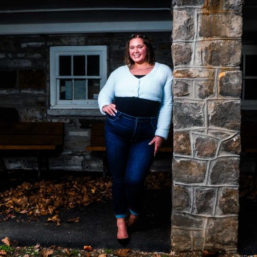 A young woman stands confidently beside a stone pillar, wearing a light sweater and jeans, with autumn leaves scattered on the ground. The setting features a rustic stone building in the background.