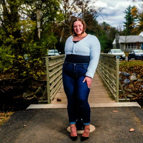 A woman stands confidently on a path in front of a wooden bridge, surrounded by trees and a residential area. She wears a light sweater and dark jeans, with heels complementing her outfit.