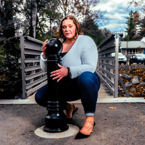 A woman crouches next to a large chess piece on a bridge, surrounded by trees and a cloudy sky. She wears a light sweater and jeans, posing confidently for the photo.