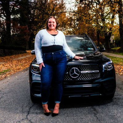 A woman stands confidently next to a black Mercedes-Benz SUV on a tree-lined street covered in autumn leaves. The scene captures a vibrant fall atmosphere.