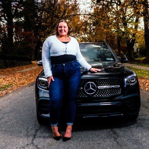 A woman stands confidently next to a black Mercedes-Benz SUV on a tree-lined street covered in autumn leaves. The scene captures a vibrant fall atmosphere.
